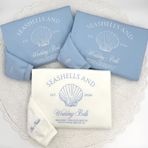May include: Three sweatshirts, two light blue and one off-white, with seashell and wedding-themed text. The text includes "SEASHELLS AND", "Wedding Bells", and "2024". The sweatshirts are arranged on a white, scalloped mat.