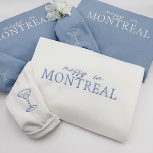 May include: Two white sweatshirts with blue embroidery that says "messy in Montreal" and a martini glass design. One sweatshirt is folded on top of the other.