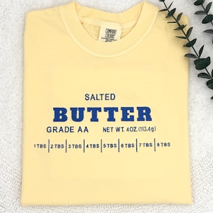May include: A yellow t-shirt with a blue graphic that reads "SALTED BUTTER GRADE AA NET WT. 4OZ. (113.4g) 1TBS 2TBS 3TBS 4TBS 5TBS 6TBS 7TBS 8TBS"