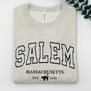 May include: A light gray heather crewneck sweatshirt featuring black embroidered text that reads SALEM MASSACHUSETTS EST. 1626, with a small black cat silhouette between the year and the abbreviation. The garment has a Jerzees brand neck label.