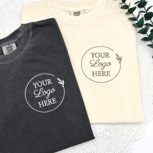 May include: Two t-shirts, one charcoal and one cream-colored, are displayed. Each shirt features a circular logo design with the text "YOUR Logo HERE" and a leaf accent. The shirts are made of a soft material and are folded neatly.
