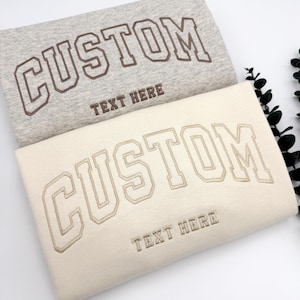 May include: Two sweatshirts with the word "CUSTOM" embroidered in a bold, outlined font. The top sweatshirt is gray with brown embroidery, and the bottom sweatshirt is beige with tan embroidery. Both sweatshirts have the text "TEXT HERE" underneath the word "CUSTOM".