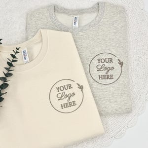 May include: Two sweatshirts, one cream-colored and one gray, are folded and displayed. Each has a circular logo that reads "YOUR Logo HERE" in a script font. The sweatshirts are made of a soft material and are likely for casual wear.