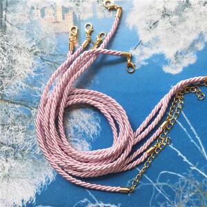 Puede incluir: Un juego de seis cordones trenzados de color rosa con cierres dorados. Los cordones están trenzados y dispuestos en forma circular.