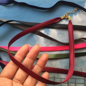 May include: A burgundy choker necklace with gold-colored chain and clasps. The choker is made of a thin, smooth ribbon. Other ribbons in various colors are visible in the background, including black, pink, and green.