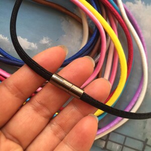 May include: A black cord necklace with a silver-colored magnetic clasp is held in a hand. Various colorful cords, including pink, yellow, and blue, are visible in the background. The necklace is simple and elegant.