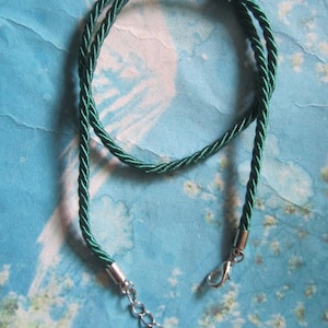 May include: A dark green rope necklace with a silver-colored clasp and chain. The rope is twisted and has a shiny appearance. The background is a light blue watercolor design.