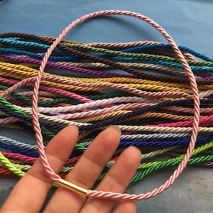 Puede incluir: Una variedad de cordones trenzados de colores, cada uno con un cierre de metal dorado, se muestran en una mano. Los cordones están dispuestos en una pila, con los extremos de los cordones superpuestos.