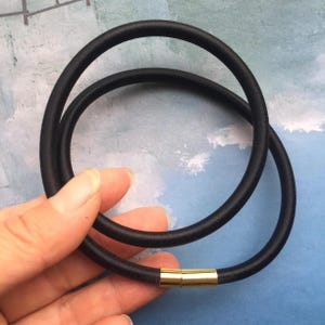 May include: Black hair tie with a gold-colored clasp. The hair tie is made of a smooth, elastic material and is designed to wrap around hair. The clasp is rectangular and connects the two ends of the tie. The background is a light blue color.