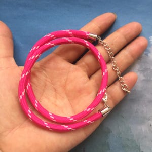 May include: A bright pink necklace with a white zigzag pattern. The necklace is made of a thick, woven material and has a silver-colored clasp and chain extender. The necklace is shown on a hand.
