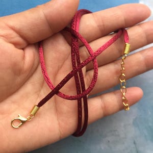 Puede incluir: Un collar de dos tonos con un cordón burdeos y rosa. El collar tiene detalles metálicos dorados, incluyendo un cierre y un extensor de cadena. El cordón está anudado en el centro.