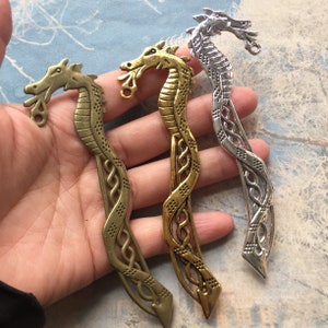 May include: Three metal dragon bookmark designs. One is silver, one is gold, and one is bronze. Each bookmark features a dragon head with intricate details and a curled tail.