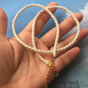 May include: Ivory-colored necklace with a twisted rope design. The necklace features a gold-colored clasp and extender chain. The rope is made of a soft, shiny material. The necklace is shown against a light blue background.