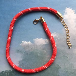 May include: Red and white patterned rope necklace with gold-colored metal accents. The necklace features a lobster clasp and an adjustable chain. The rope is thick and has a repeating white pattern.