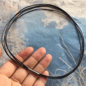 Può includere: Una collana in cordoncino di cuoio nero con una chiusura in metallo argentato. Il cordoncino è a strati e sembra regolabile.