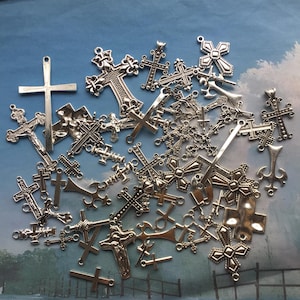 Könnte beinhalten: Eine Sammlung silberfarbener Kreuze in verschiedenen Größen und Designs. Einige Kreuze weisen aufwendige Details auf, andere sind schlicht. Die Kreuze sind vor einem blau-weißen Hintergrund verstreut und erzeugen eine visuell interessante Darstellung.