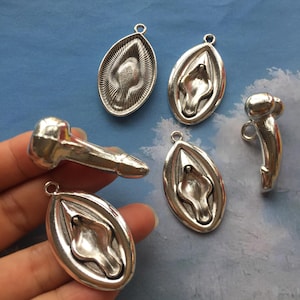 May include: Silver-toned metal pendants in various shapes. The collection includes oval pendants with detailed designs and phallic shapes. The pendants are likely for jewelry making or crafting.