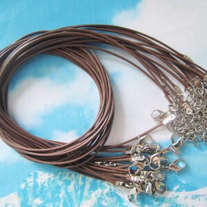 Puede incluir: Un conjunto de cordones de algodón encerado marrón con cierres de langosta plateados. Los cordones están dispuestos en un círculo con los cierres en el exterior.