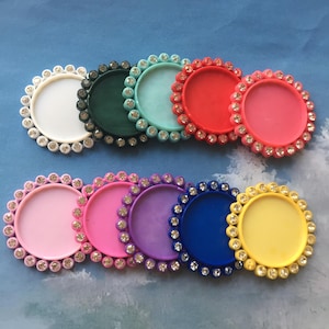 May include: Assortment of ten round, flat, plastic, blank,  cabochons with a rhinestone border. The cabochons are in various colours: white, green, blue, red, pink, light pink, purple, blue, yellow.