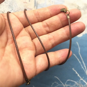 Puede incluir: Un collar de cordón encerado marrón con un cierre y un anillo de salto de bronce.