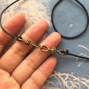 Puede incluir: Primer plano de un collar de cordón negro con un cierre de latón. El cierre tiene un diseño decorativo con un pequeño círculo en el centro.