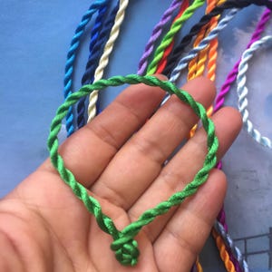 Könnte beinhalten: Ein grünes geflochtenes Kordel-Armband mit einem Knoten am Ende. Das Armband wird in einer Hand gehalten, und im Hintergrund befinden sich weitere geflochtene Kordeln in verschiedenen Farben.