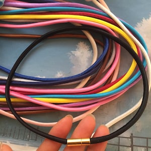 May include: A black necklace with a gold clasp is held in front of a collection of colorful cords. The cords are in various colors, including pink, blue, yellow, and white. The necklace appears to be made of a smooth, round material.