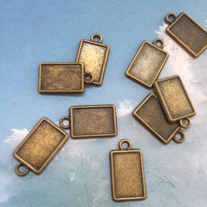 Puede incluir: Diez colgantes rectangulares de metal dorado antiguo con un bucle en la parte superior. Los colgantes están en blanco y listos para ser personalizados.