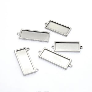 Puede incluir: Cinco bandejas colgantes rectangulares de metal plateado con bordes elevados y pequeños bucles circulares en los extremos. Estos accesorios de joyería cuentan con una superficie interior lisa y plana para colocar cabujones.