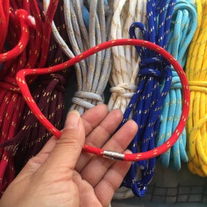 May include: A red rope necklace with a silver clasp is displayed against a backdrop of various coloured ropes. The ropes are in shades of red, grey, white, blue, and yellow, each with a gold metallic thread.