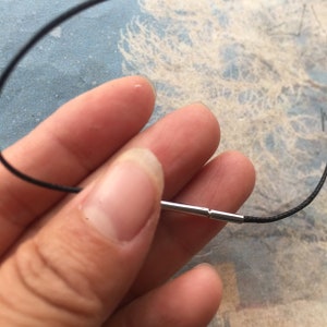 Pode incluir: Close-up de uma ponta de cordão de metal prateado com um cordão preto preso. A ponta do cordão é segurada por uma mão.