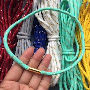 May include: A close-up of a light green necklace with a gold clasp, displayed in front of bundles of colorful cords. The cords are in various colors, including red, gray, blue, white, mint green, and yellow, all with gold metallic accents.