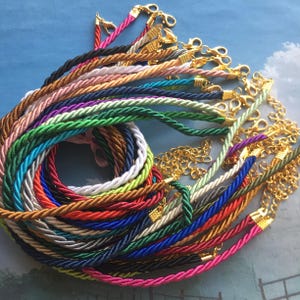 May include: A collection of colorful twisted rope necklaces with gold-colored clasps and chain extenders. The ropes are in various colors, including pink, green, blue, and brown. The necklaces are arranged in a circular pattern against a light blue background.