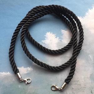 May include: Black twisted rope strap with silver-tone metal clasps. The rope is tightly wound, creating a textured surface. The clasps are attached to the ends of the rope, ready for use. The strap is designed for bags or other accessories.