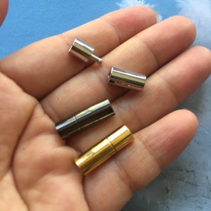 Puede incluir: Colección de cuatro cierres de joyería metálicos en plata, plata oscura y oro. Los cierres son cilíndricos y magnéticos, diseñados para una fácil fijación. Un cierre tiene un pequeño poste. Los cierres se muestran sobre un fondo azul claro.
