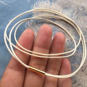 Könnte beinhalten: Eine weiße Lederkordelkette mit einem goldenen Verschluss. Die Kordel ist geschichtet und scheint verstellbar zu sein.