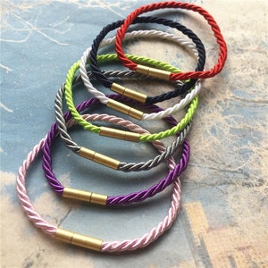 May include: A set of eight colorful braided rope bracelets with gold metal clasps. The bracelets are stacked on top of each other, with the red bracelet on top and the pink bracelet on the bottom.