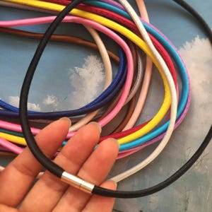 May include: A black necklace with a silver clasp is held in a hand. The necklace is surrounded by a variety of colorful necklaces in shades of pink, yellow, blue, and white. The necklaces appear to be made of a smooth, cord-like material.