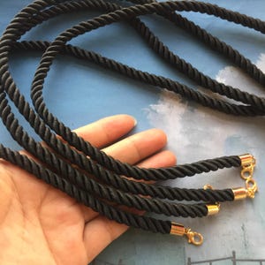 May include: Three black braided cords with gold metal clasps at the ends. The cords are twisted together and are lying on a hand.