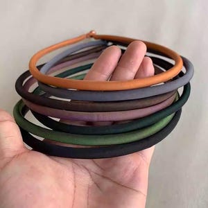 May include: A collection of colorful, round hair ties in various shades, including orange, gray, green, and black. The hair ties are stacked in a hand, showcasing the range of colors and textures. The ties appear to be made of a soft, fabric-covered material.