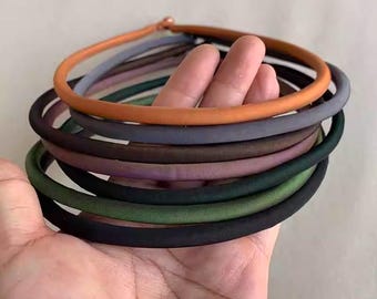 May include: A collection of colorful, round hair ties in various shades, including orange, gray, green, and black. The hair ties are stacked in a hand, showcasing the range of colors and textures. The ties appear to be made of a soft, fabric-covered material.