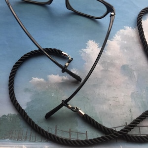 Puede incluir: Gafas negras con una cadena de gafas de estilo cuerda negra. La cadena está unida a las patillas de las gafas con cierres de metal plateado.