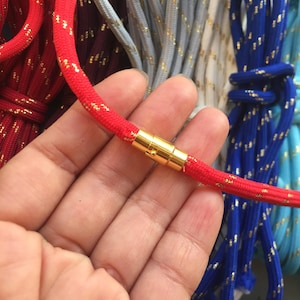 May include: A close-up of a red rope with gold flecks and a gold clasp. The rope is held in a hand, with other colorful ropes visible in the background. The rope is approximately 0.5 inches in diameter.