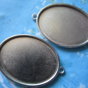 Puede incluir: Dos blancos ovalados de metal de color plateado con una superficie lisa ligeramente texturizada. Los blancos tienen un pequeño bucle en un lado para sujetar a una cadena o cordón.