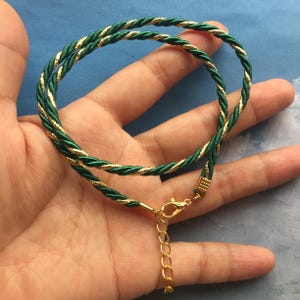 May include: A green and gold rope necklace with a gold clasp. The necklace is made of a braided rope with a gold thread running through it.