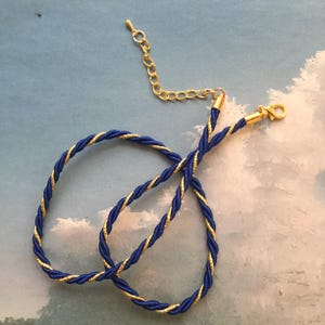 May include: A blue and gold twisted cord necklace with a gold-colored clasp and chain. The necklace is made of a twisted blue cord with a gold metallic thread woven throughout. The clasp and chain are gold-colored.
