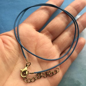 Puede incluir: Un collar de cordón azul con un cierre dorado. El cordón está doblado y el cierre está unido a una cadena pequeña.