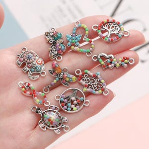 May include: Assortment of colourful charm pendants with various designs, including a butterfly, elephant, dragonfly, tree, hand, heart, infinity symbol, and turtle. Each charm is made of silver-toned metal and features colourful enamel details.