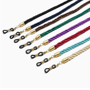 May include: A set of eight eyeglass chains with gold-tone clasps and black eyeglass holders. The chains are made of braided rope in various colors, including black, navy blue, brown, green, purple, red, turquoise, and cream.