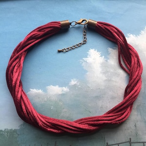 Puede incluir: Un collar de cordón de satén rojo con un cierre de latón. El collar está hecho de múltiples hebras de cordón de satén que están trenzadas juntas.